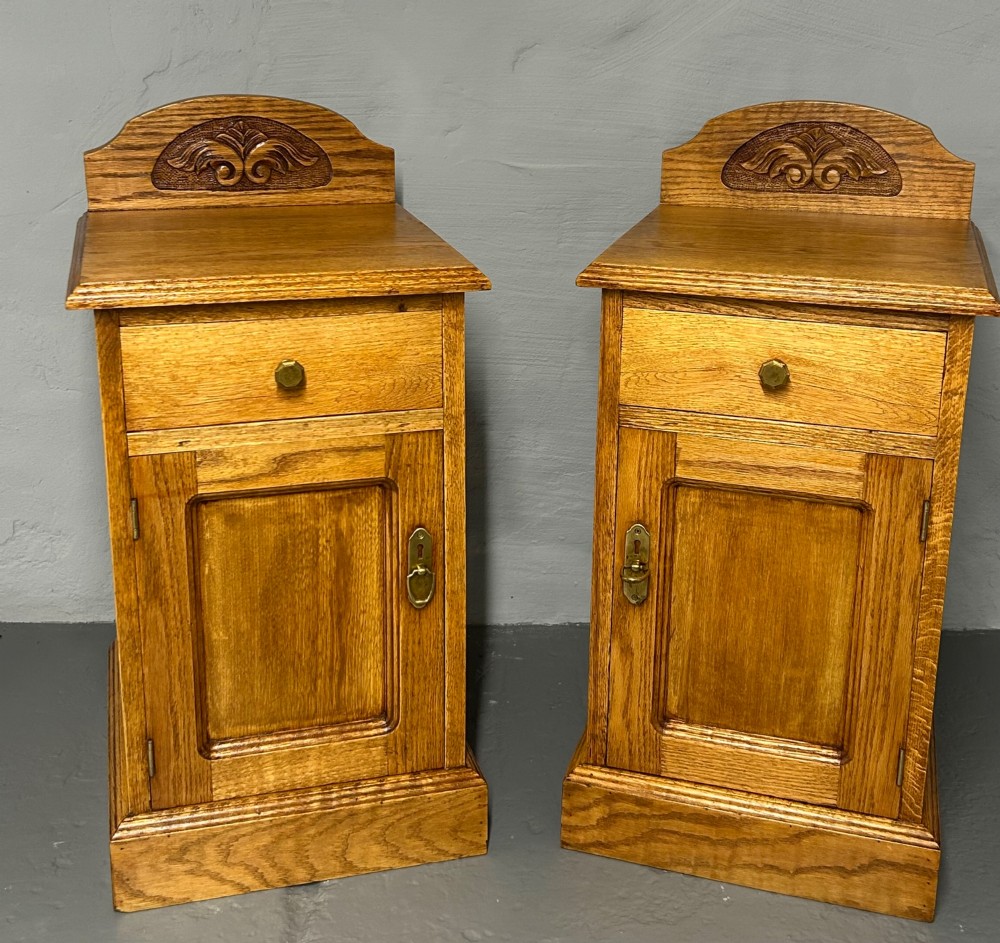 A matching pair of light oak bedside cabinets featuring carved crest galleries, single drawers, single panel doors, and brass fittings.