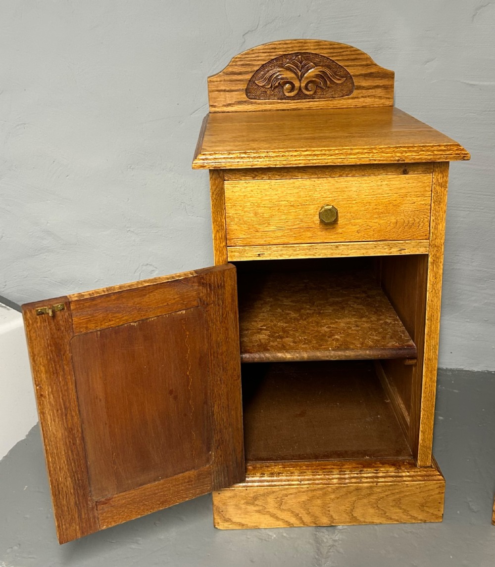 A matching pair of light oak bedside cabinets featuring carved crest galleries, single drawers, single panel doors, and brass fittings. View III