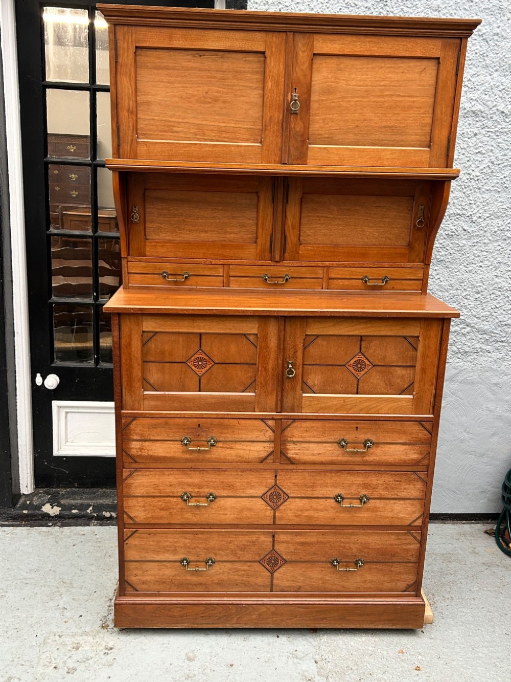 A tall three-tier Aesthetic Movement wooden cabinet featuring geometric diamond wood inlay, panelled doors, and drawers with brass drop handles. View II