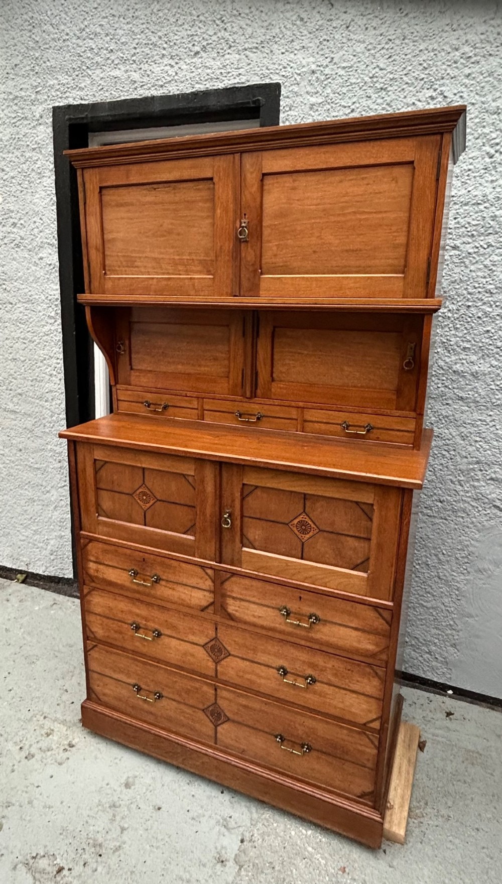 A tall three-tier Aesthetic Movement wooden cabinet featuring geometric diamond wood inlay, panelled doors, and drawers with brass drop handles. View III