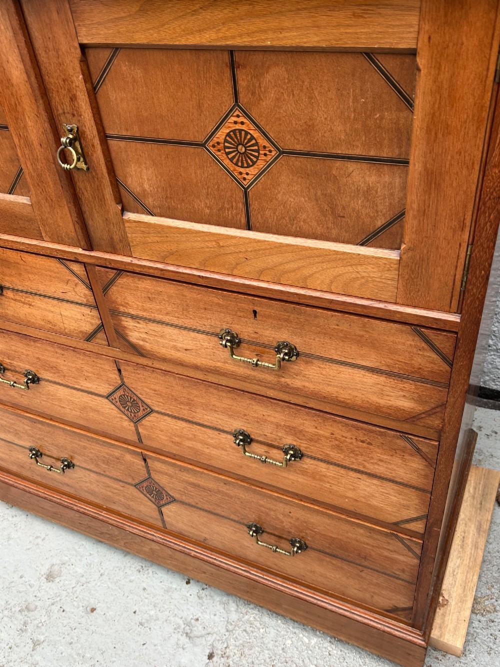 A tall three-tier Aesthetic Movement wooden cabinet featuring geometric diamond wood inlay, panelled doors, and drawers with brass drop handles. View IV