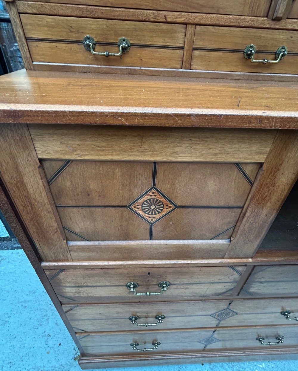 A tall three-tier Aesthetic Movement wooden cabinet featuring geometric diamond wood inlay, panelled doors, and drawers with brass drop handles. View V