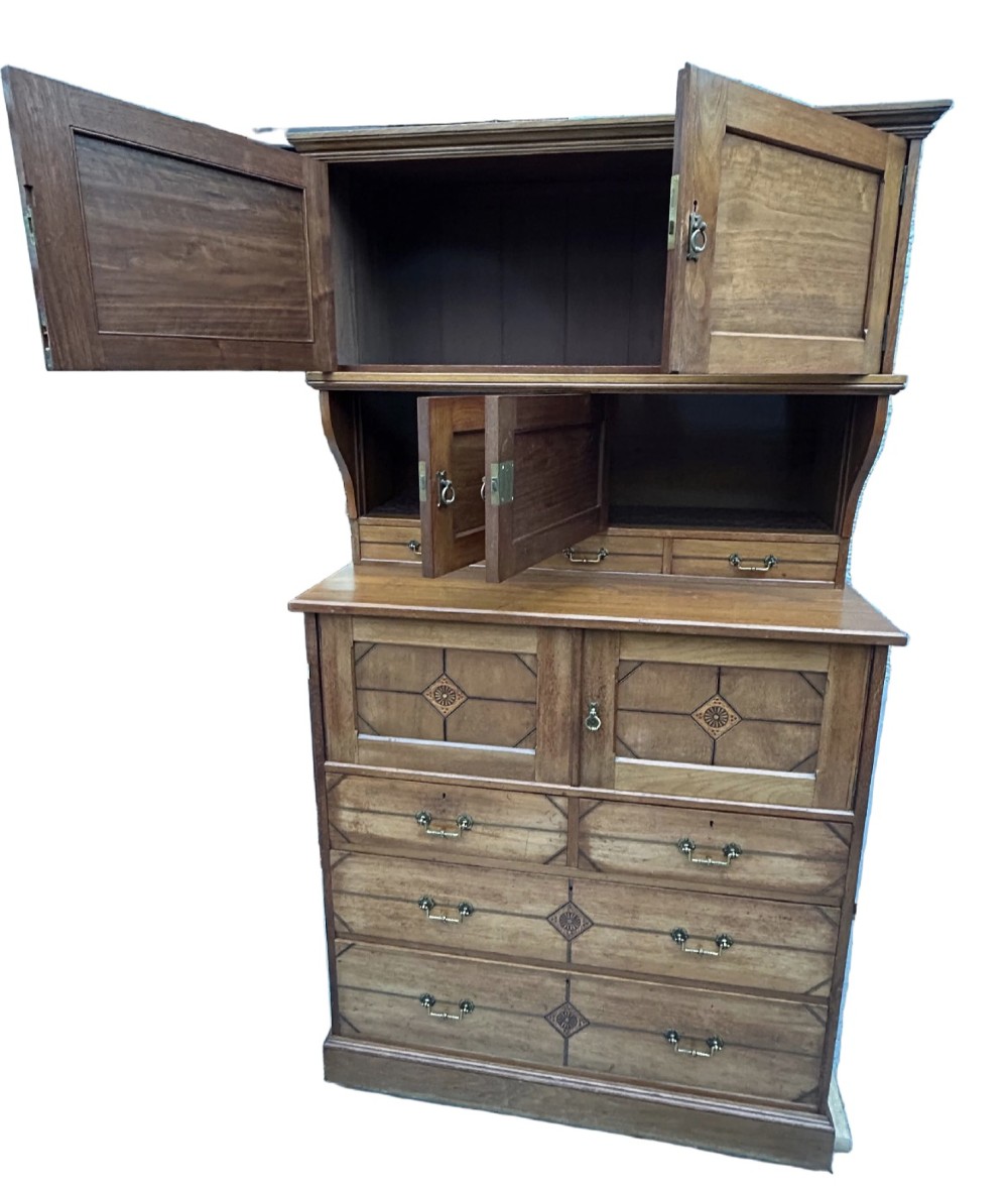 A tall three-tier Aesthetic Movement wooden cabinet featuring geometric diamond wood inlay, panelled doors, and drawers with brass drop handles. View VII