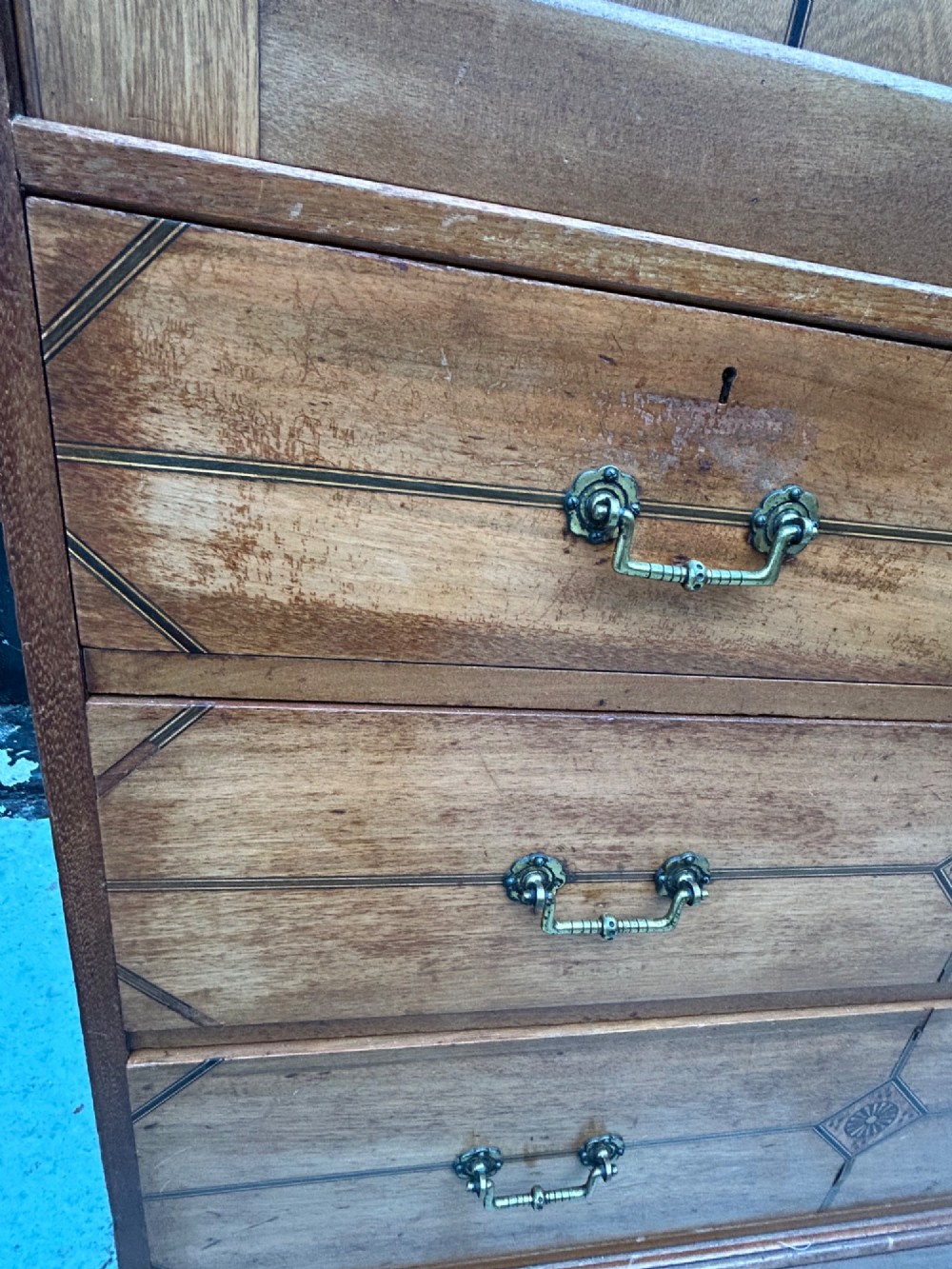 A tall three-tier Aesthetic Movement wooden cabinet featuring geometric diamond wood inlay, panelled doors, and drawers with brass drop handles. View VIII