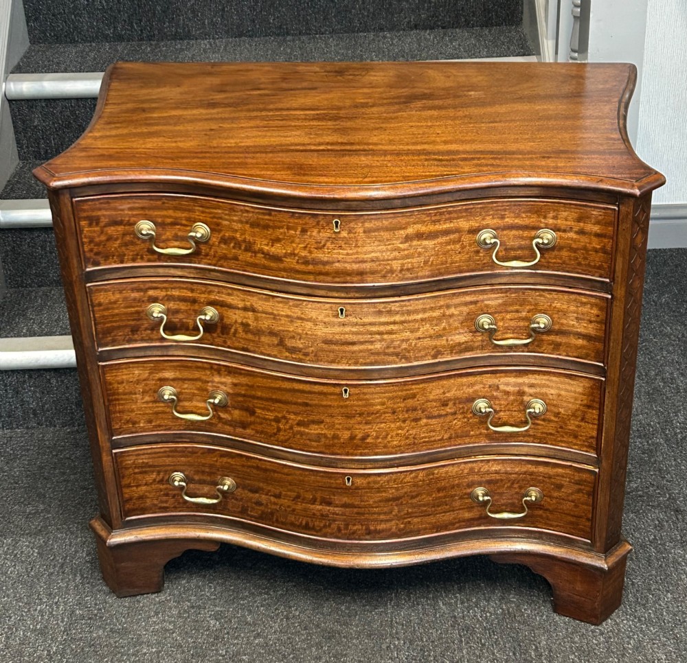 A Georgian Revival mahogany chest of drawers featuring a serpentine front, four graduated drawers with brass handles, blind fretwork on canted corners, and bracket feet.