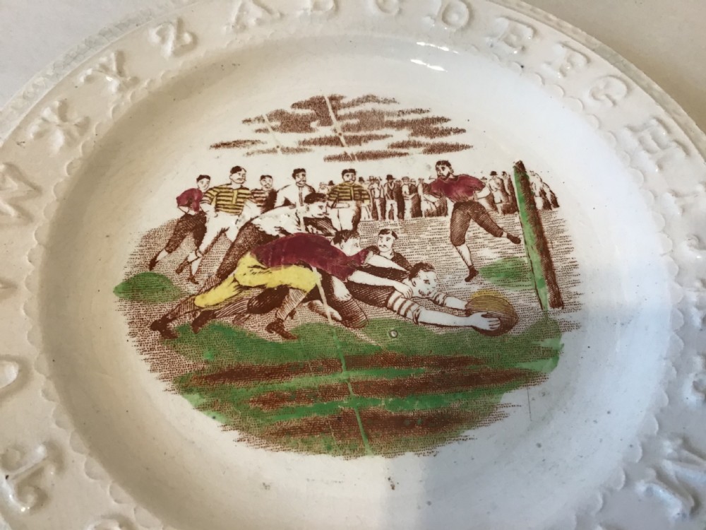 A circular cream earthenware nursery plate featuring an embossed alphabet rim surrounding a transfer-printed central scene depicting a rugby match. View II
