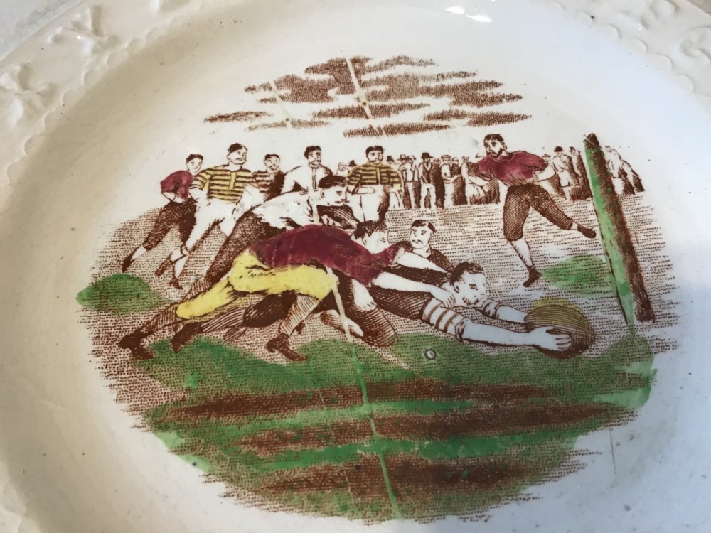 A circular cream earthenware nursery plate featuring an embossed alphabet rim surrounding a transfer-printed central scene depicting a rugby match. View IV