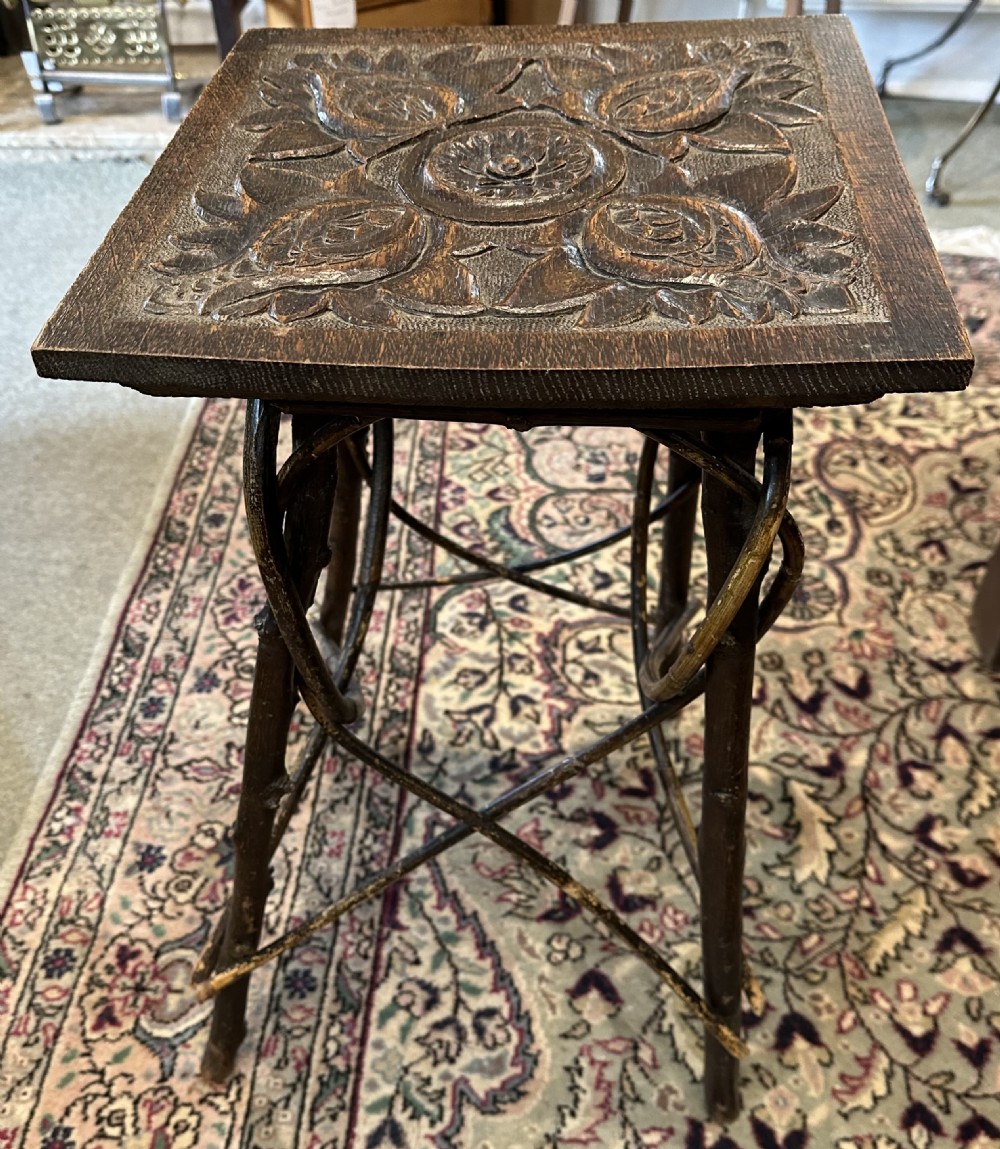 A dark brown wooden side table featuring a square top with relief floral carving supported by a rustic twig base.