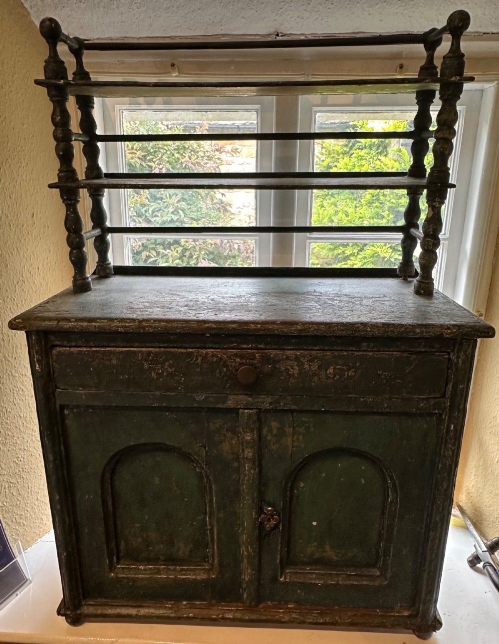 An antique French dark green painted miniature dresser featuring a tiered spindle plate rack above a single drawer and twin arched panel cupboard doors.