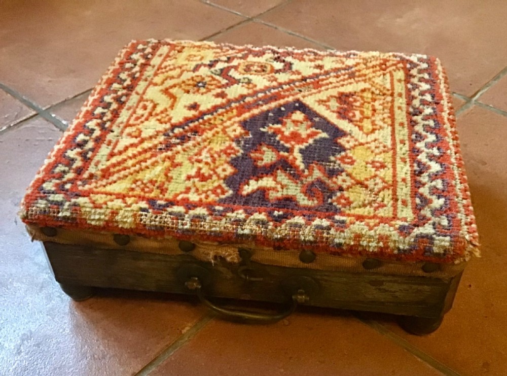 An antique Victorian carriage foot warmer featuring a square patterned carpet top with stud detailing, a wooden base with turned feet, and a metal carry handle.