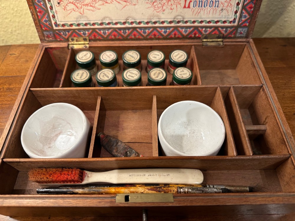 An open Victorian wooden artist box containing glass pigment jars, ceramic bowls, a brush, and an illustrated lid label for George Rowney and Company. View III