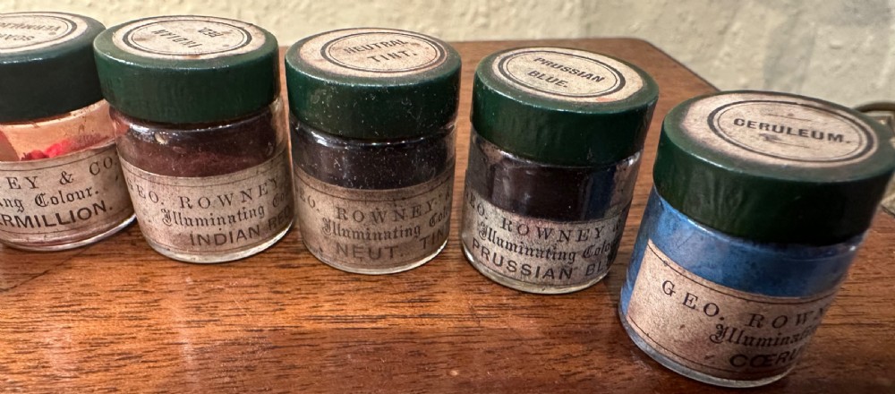 An open Victorian wooden artist box containing glass pigment jars, ceramic bowls, a brush, and an illustrated lid label for George Rowney and Company. View VI