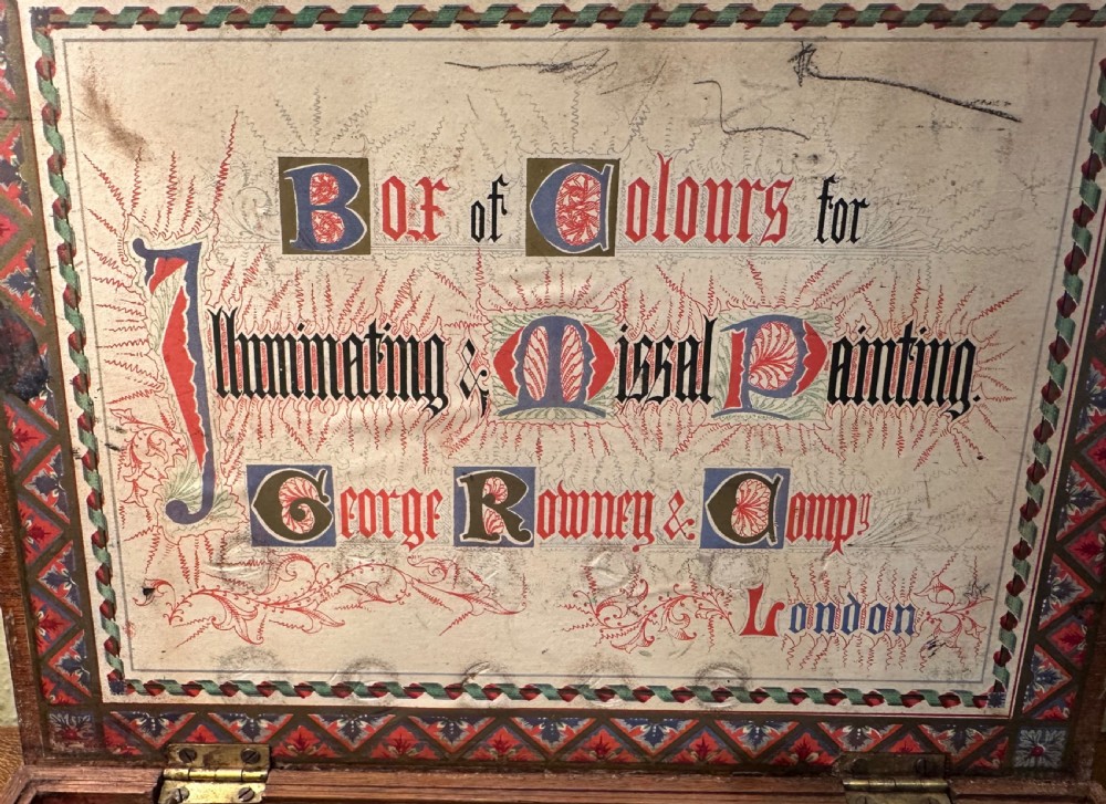 An open Victorian wooden artist box containing glass pigment jars, ceramic bowls, a brush, and an illustrated lid label for George Rowney and Company. View VII