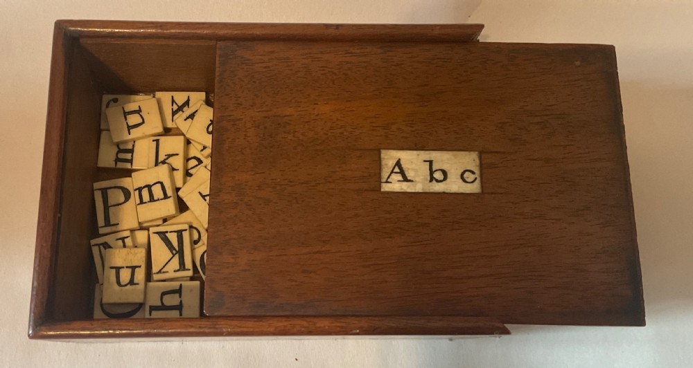A mahogany box with a sliding lid featuring an inlaid bone plaque inscribed A b c, containing rectangular bone tiles stamped with black lettering.