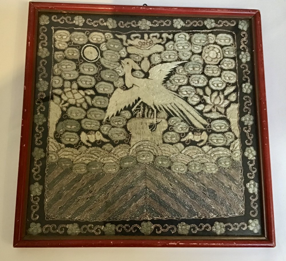 A square framed Chinese silk rank badge featuring a central embroidered bird surrounded by clouds, bats, and water waves in cream silk thread against a dark background.