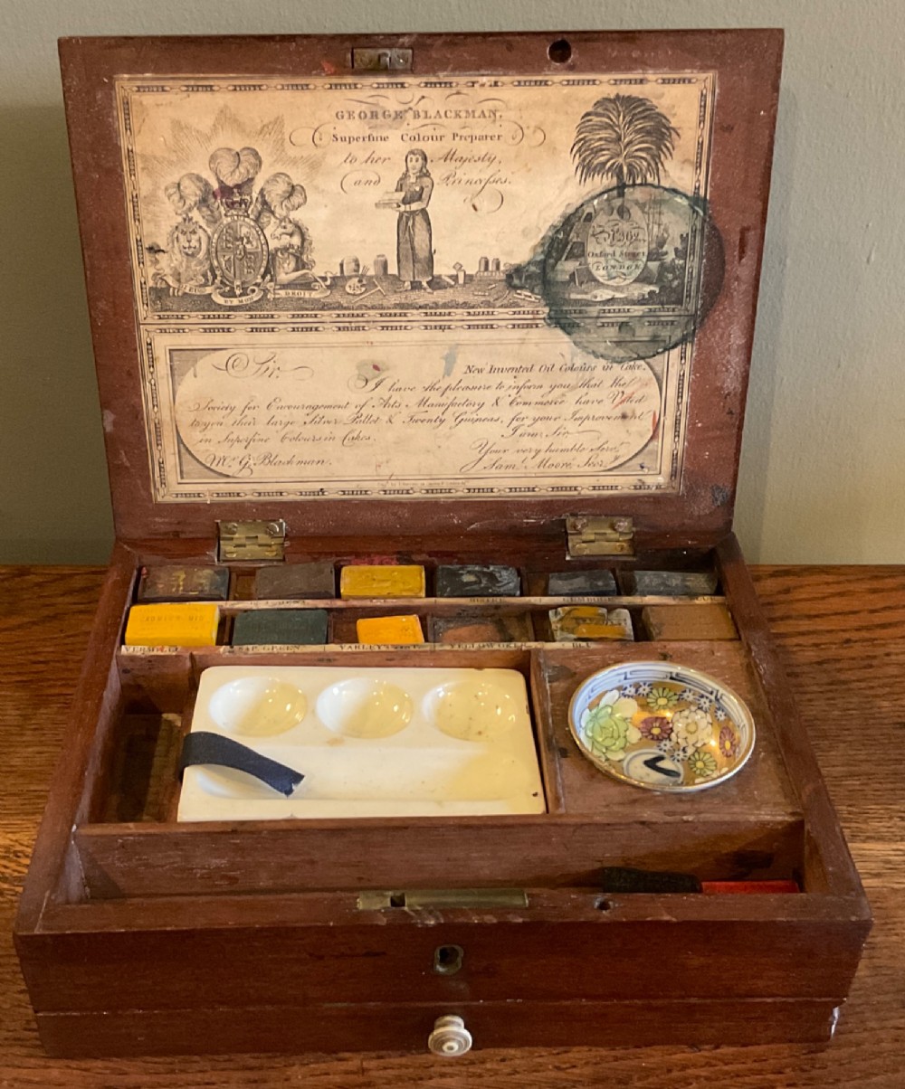 An open Georgian mahogany artist paintbox displaying original pigment cakes, a porcelain palette, a decorated ceramic dish, and a printed maker label inside the lid.