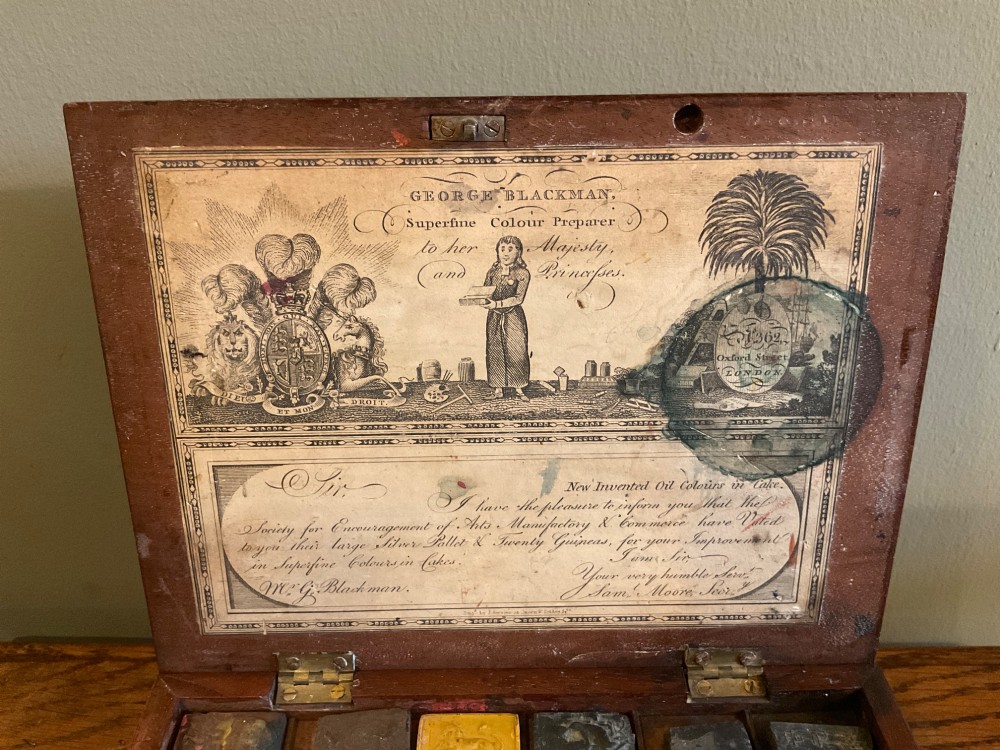 An open Georgian mahogany artist paintbox displaying original pigment cakes, a porcelain palette, a decorated ceramic dish, and a printed maker label inside the lid. View II
