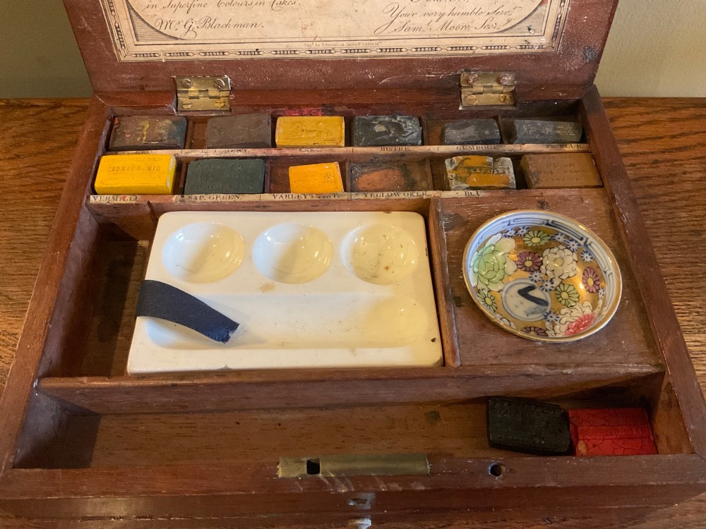 An open Georgian mahogany artist paintbox displaying original pigment cakes, a porcelain palette, a decorated ceramic dish, and a printed maker label inside the lid. View III