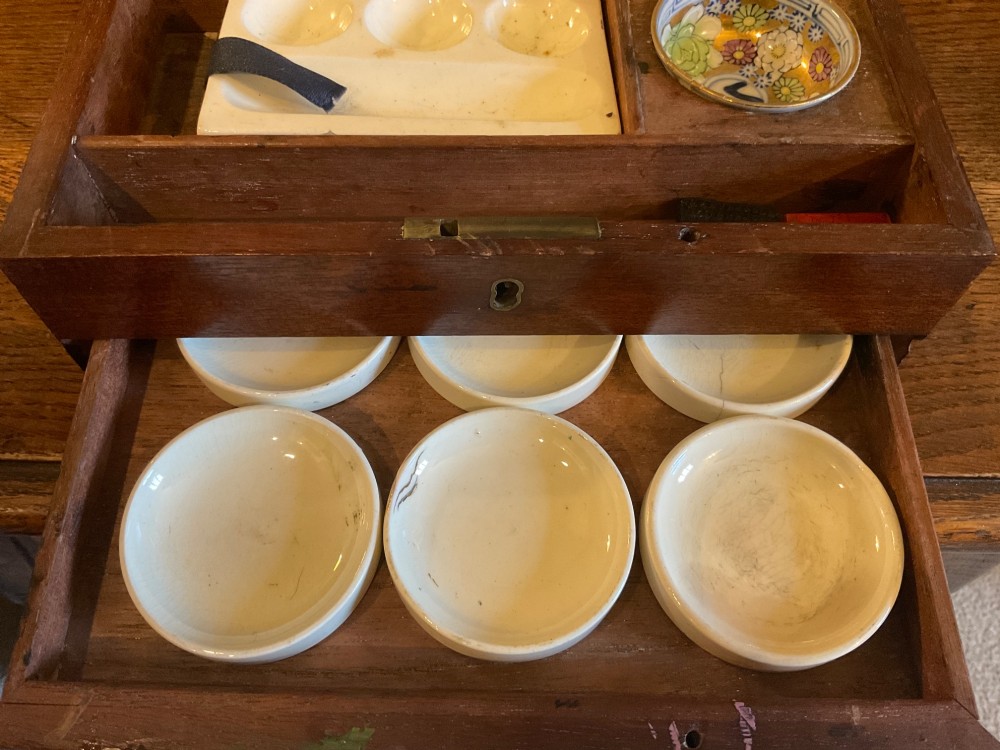 An open Georgian mahogany artist paintbox displaying original pigment cakes, a porcelain palette, a decorated ceramic dish, and a printed maker label inside the lid. View IV