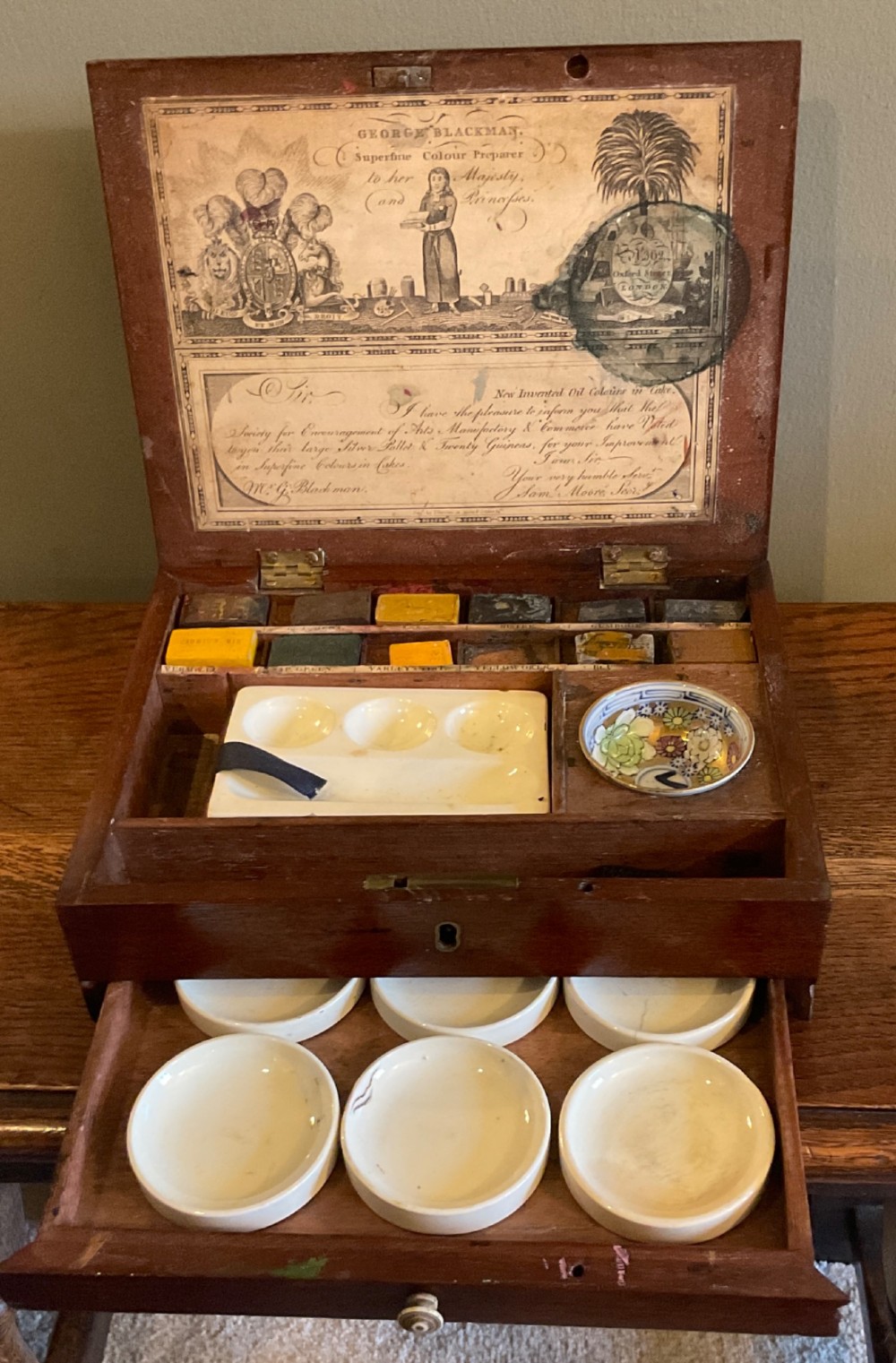 An open Georgian mahogany artist paintbox displaying original pigment cakes, a porcelain palette, a decorated ceramic dish, and a printed maker label inside the lid. View V