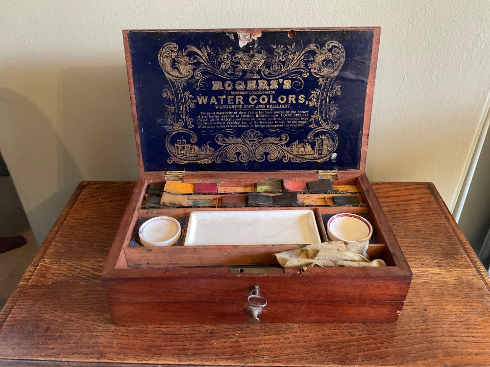 An open Victorian mahogany artist box featuring a gold-printed trade label inside the lid, fitted compartments containing watercolour blocks, and ceramic mixing dishes.