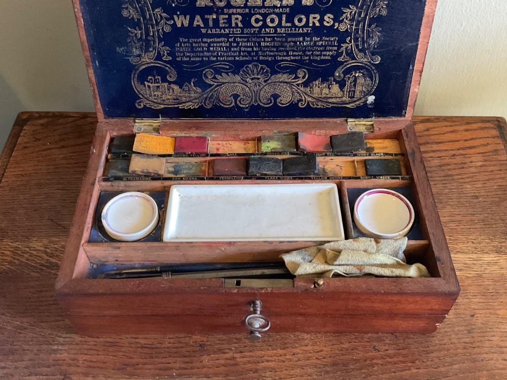 An open Victorian mahogany artist box featuring a gold-printed trade label inside the lid, fitted compartments containing watercolour blocks, and ceramic mixing dishes. View IV