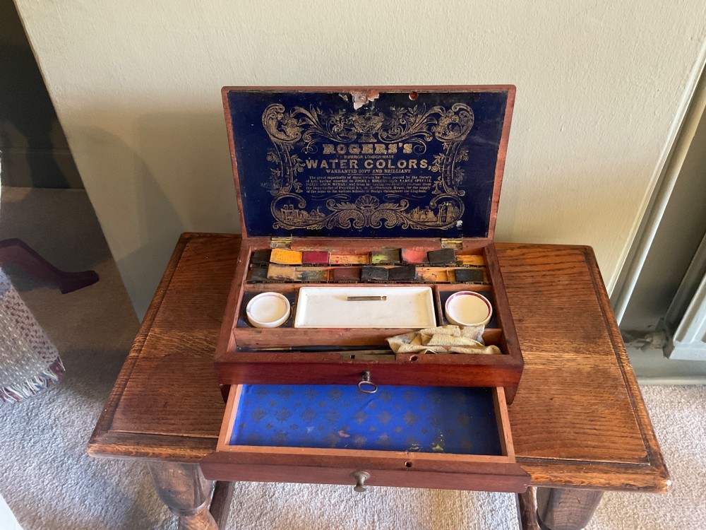 An open Victorian mahogany artist box featuring a gold-printed trade label inside the lid, fitted compartments containing watercolour blocks, and ceramic mixing dishes. View V