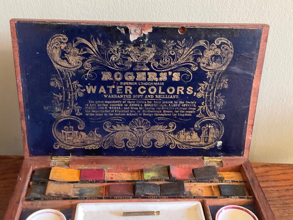 An open Victorian mahogany artist box featuring a gold-printed trade label inside the lid, fitted compartments containing watercolour blocks, and ceramic mixing dishes. View VI