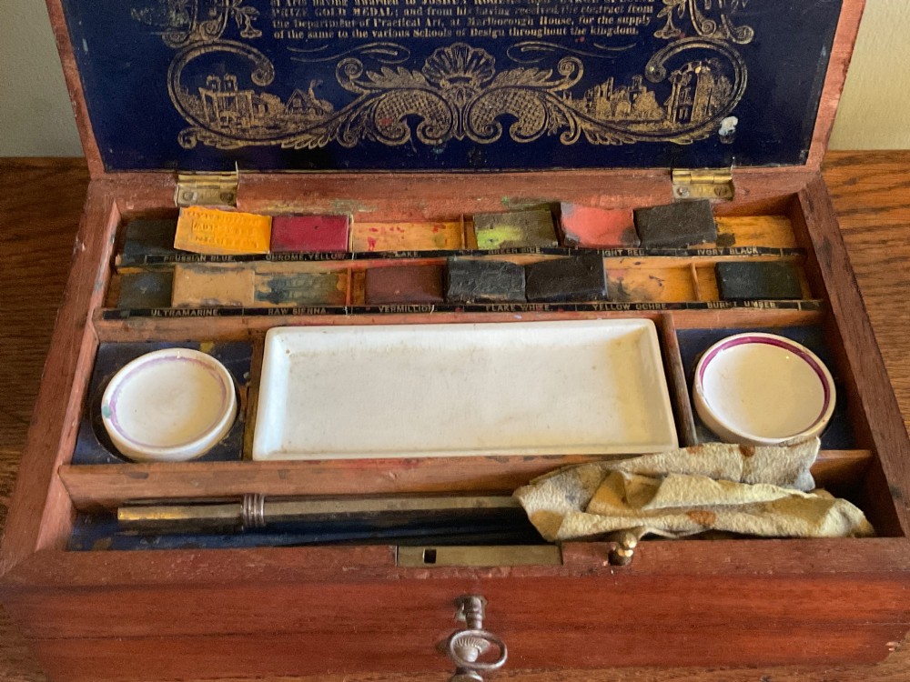 An open Victorian mahogany artist box featuring a gold-printed trade label inside the lid, fitted compartments containing watercolour blocks, and ceramic mixing dishes. View VII