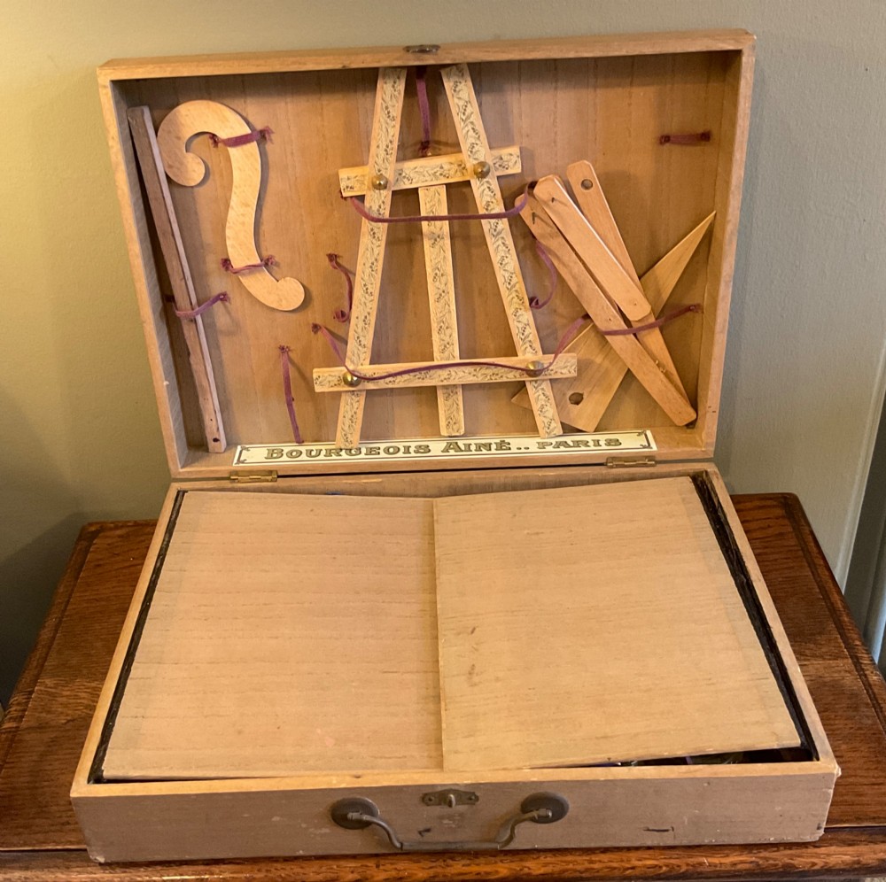 An open French beechwood artist paintbox displaying miniature wood easels, set squares, and velvet-lined compartments containing paint cakes and crayons. View III