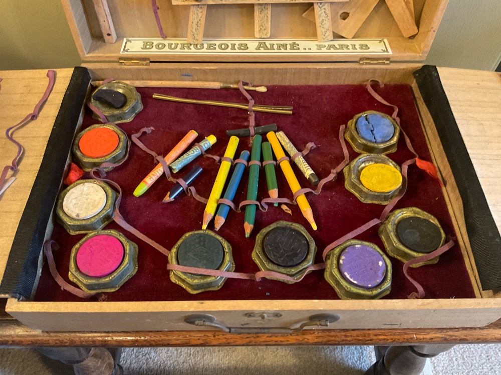 An open French beechwood artist paintbox displaying miniature wood easels, set squares, and velvet-lined compartments containing paint cakes and crayons. View IV