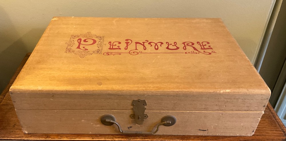 An open French beechwood artist paintbox displaying miniature wood easels, set squares, and velvet-lined compartments containing paint cakes and crayons. View V