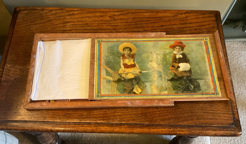 An antique wooden box lid featuring a colourful lithographed paper illustration of two children seated outdoors within a geometric border. View II