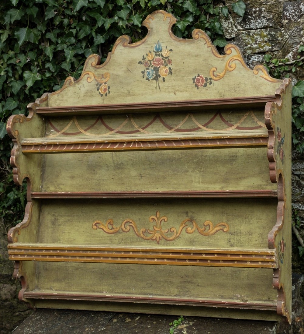 A sage green hand-painted wooden wall shelf unit featuring a decorative scalloped crest with floral motifs and three open display shelves with front retaining bars.