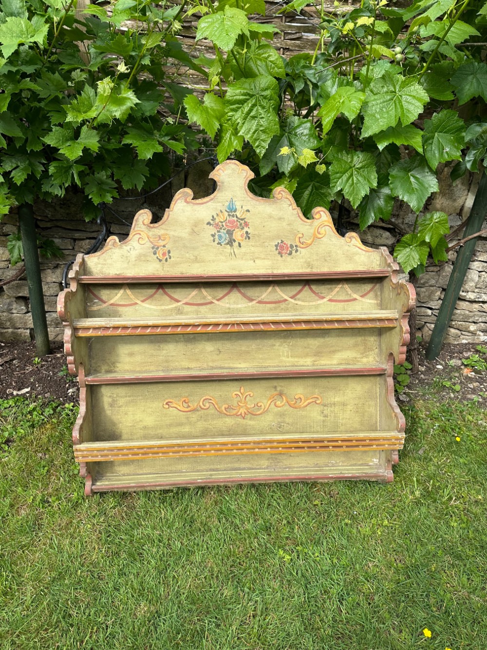 A sage green hand-painted wooden wall shelf unit featuring a decorative scalloped crest with floral motifs and three open display shelves with front retaining bars. View II