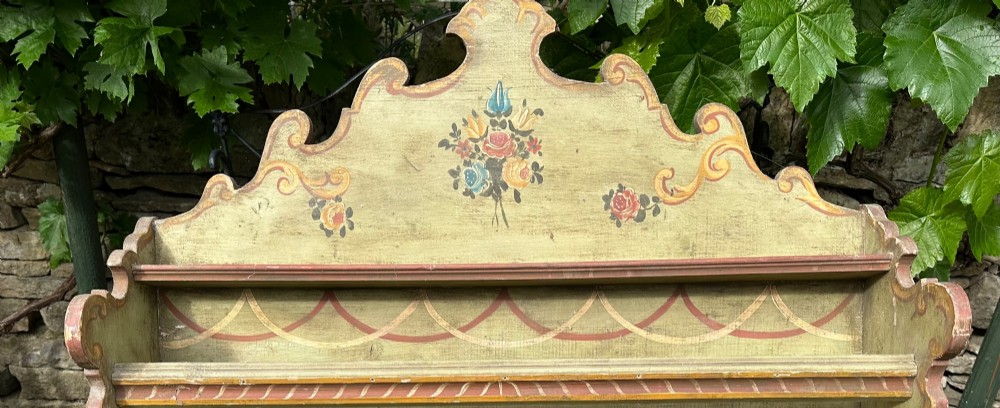 A sage green hand-painted wooden wall shelf unit featuring a decorative scalloped crest with floral motifs and three open display shelves with front retaining bars. View IV