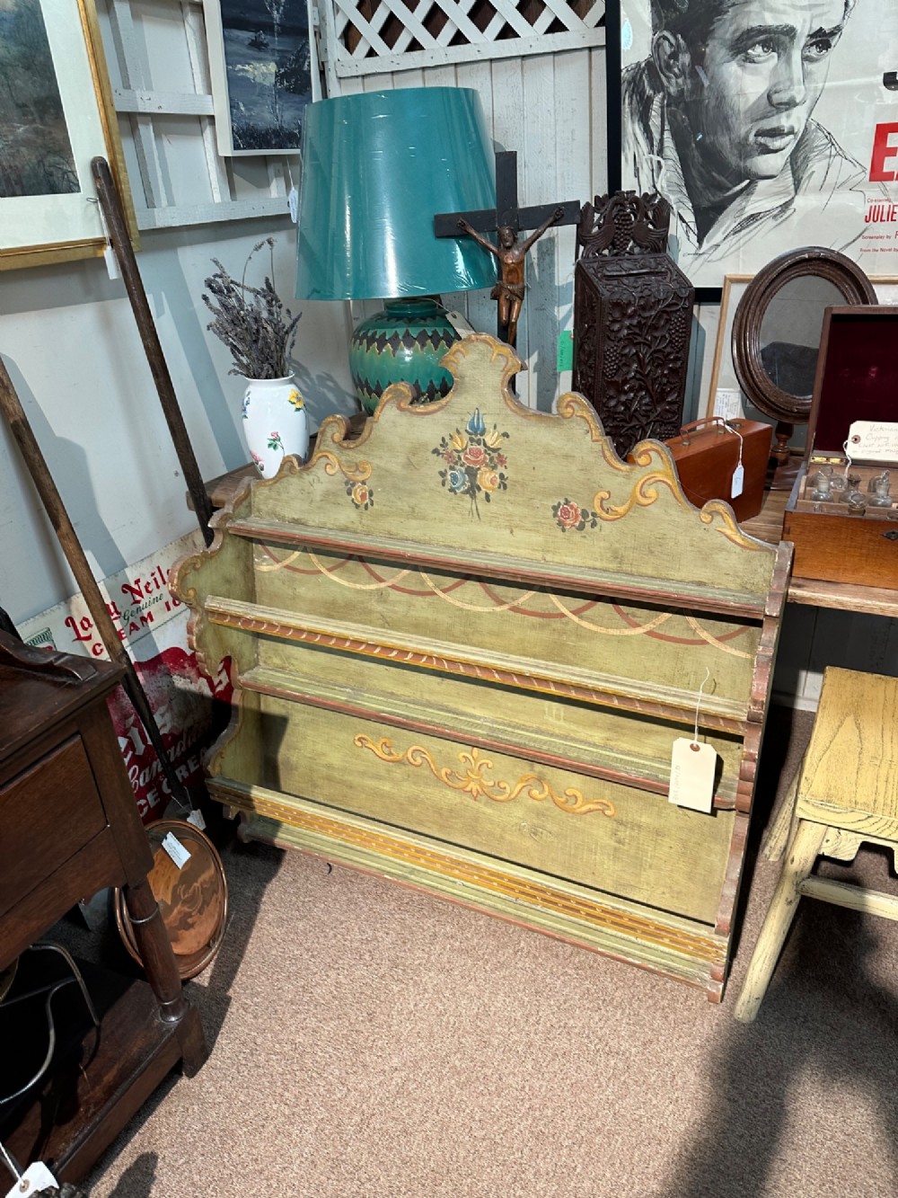 A sage green hand-painted wooden wall shelf unit featuring a decorative scalloped crest with floral motifs and three open display shelves with front retaining bars. View V