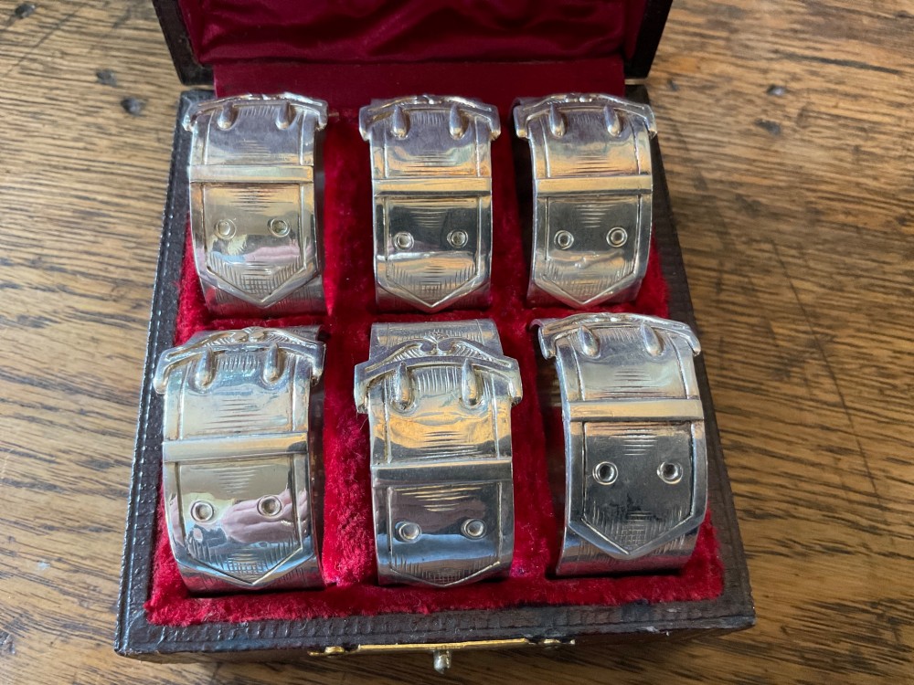 A set of six silver napkin rings styled like belt buckles presented inside a red velvet-lined presentation case.