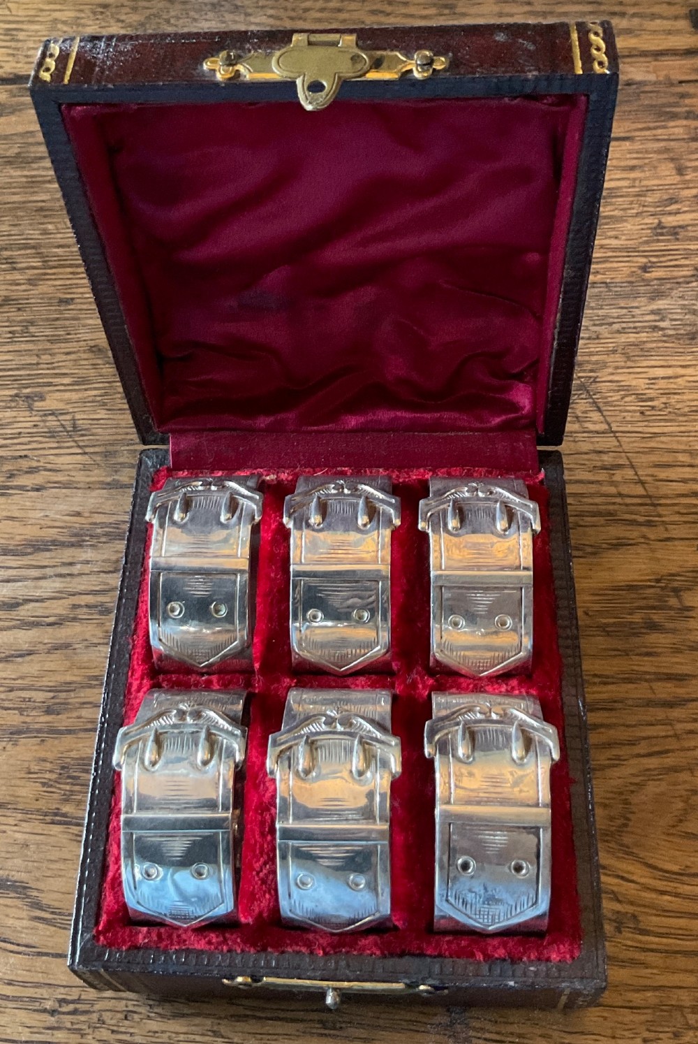 A set of six silver napkin rings styled like belt buckles presented inside a red velvet-lined presentation case. View II