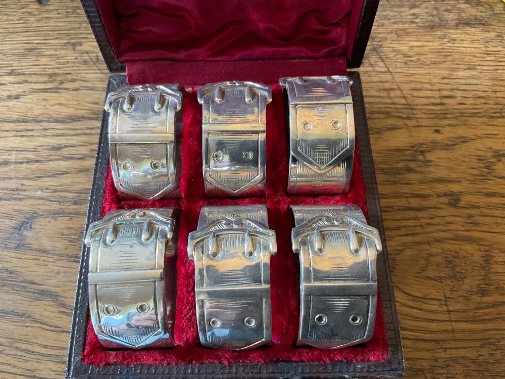 A set of six silver napkin rings styled like belt buckles presented inside a red velvet-lined presentation case. View VI