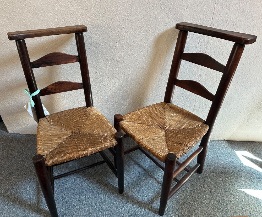 A pair of dark oak antique chapel chairs featuring woven rush seats, ladder back splats, and integrated top book ledges.