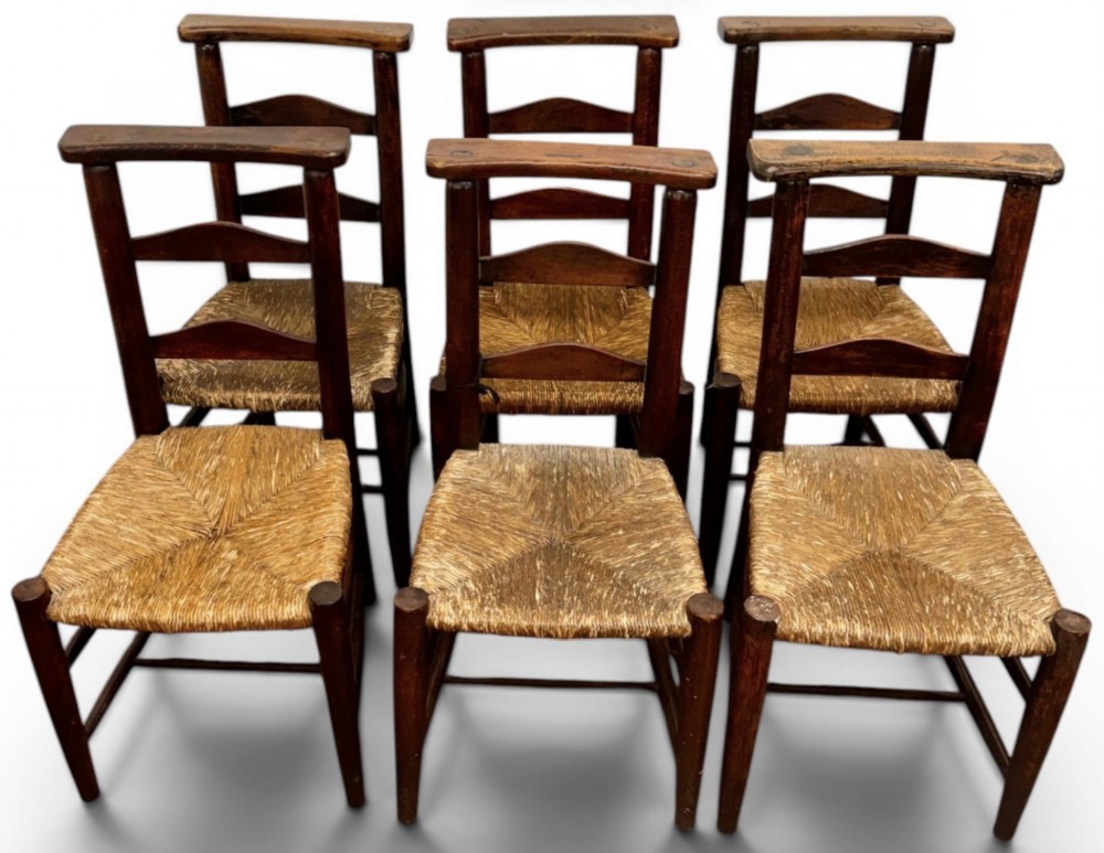 A pair of dark oak antique chapel chairs featuring woven rush seats, ladder back splats, and integrated top book ledges. View X