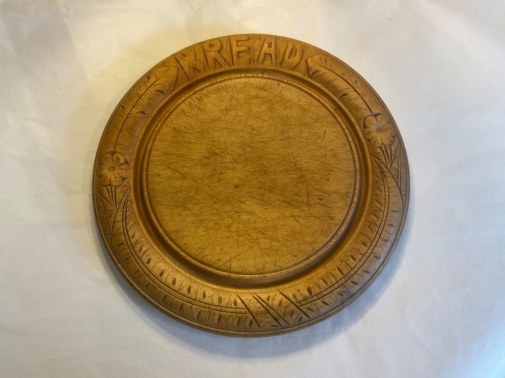A circular antique carved wooden bread board featuring the word BREAD in relief alongside carved floral motifs around the border and knife marks in the centre.