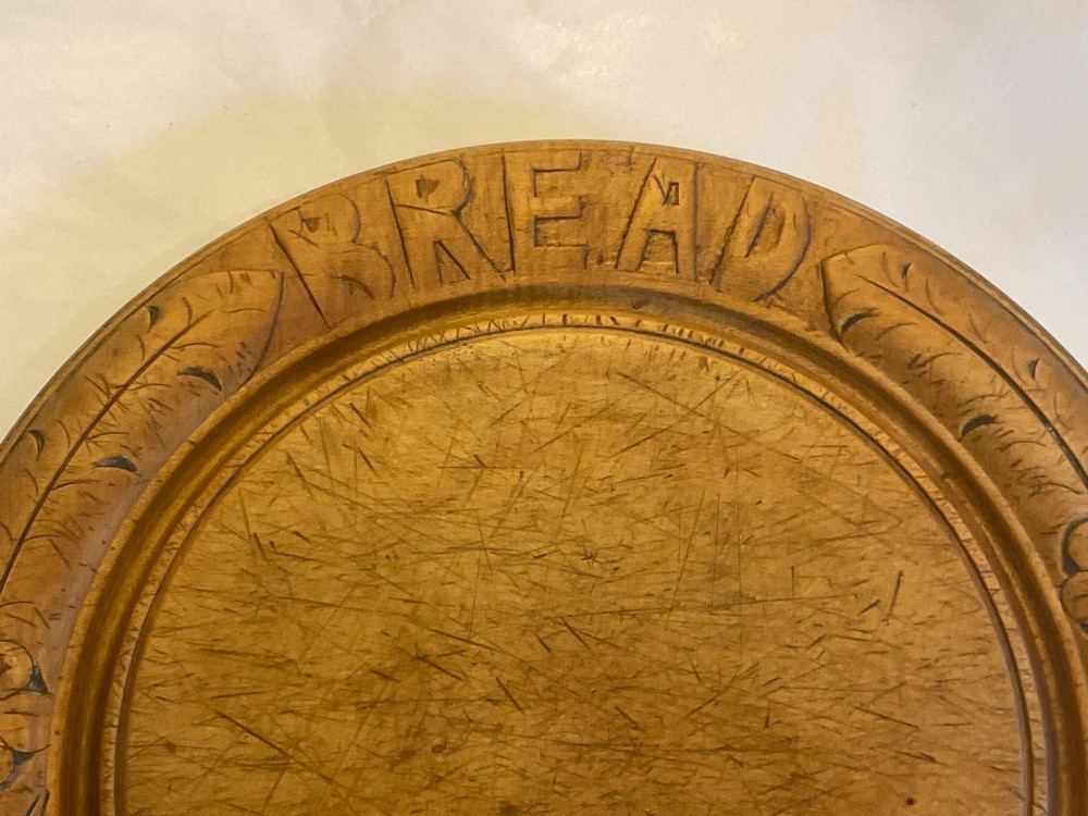 A circular antique carved wooden bread board featuring the word BREAD in relief alongside carved floral motifs around the border and knife marks in the centre. View II