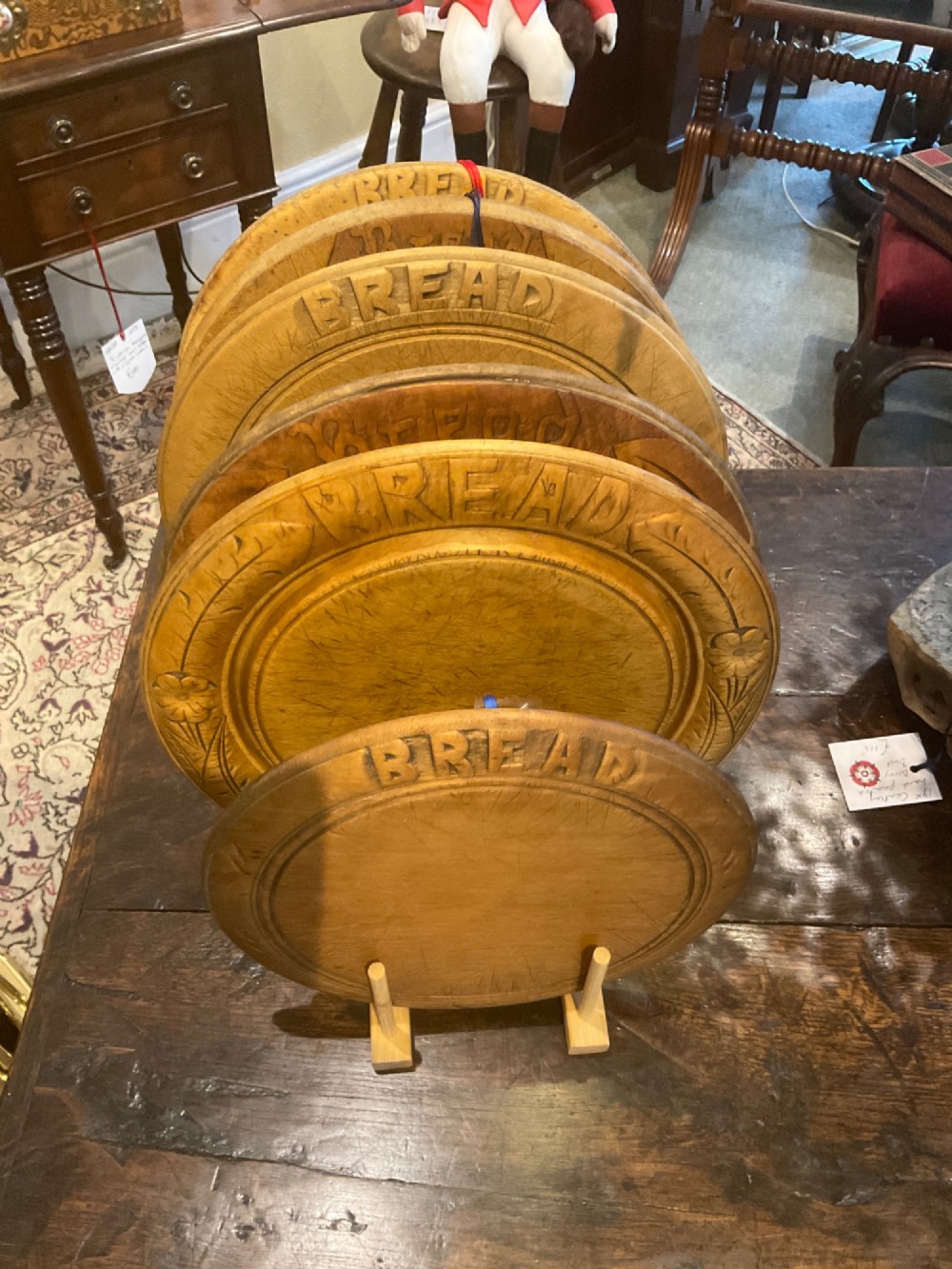 A circular antique carved wooden bread board featuring the word BREAD in relief alongside carved floral motifs around the border and knife marks in the centre. View VII