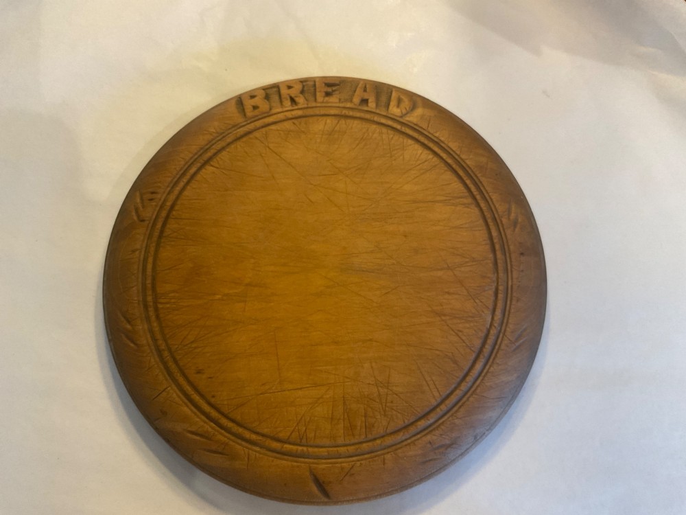 A circular antique wooden bread board featuring hand-carved lettering reading BREAD along the upper rim and knife marks across the centre.