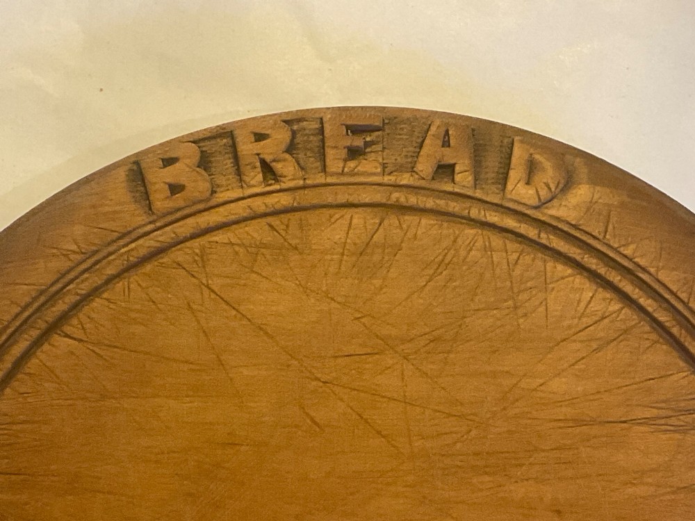 A circular antique wooden bread board featuring hand-carved lettering reading BREAD along the upper rim and knife marks across the centre. View II