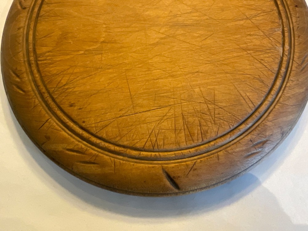 A circular antique wooden bread board featuring hand-carved lettering reading BREAD along the upper rim and knife marks across the centre. View IV