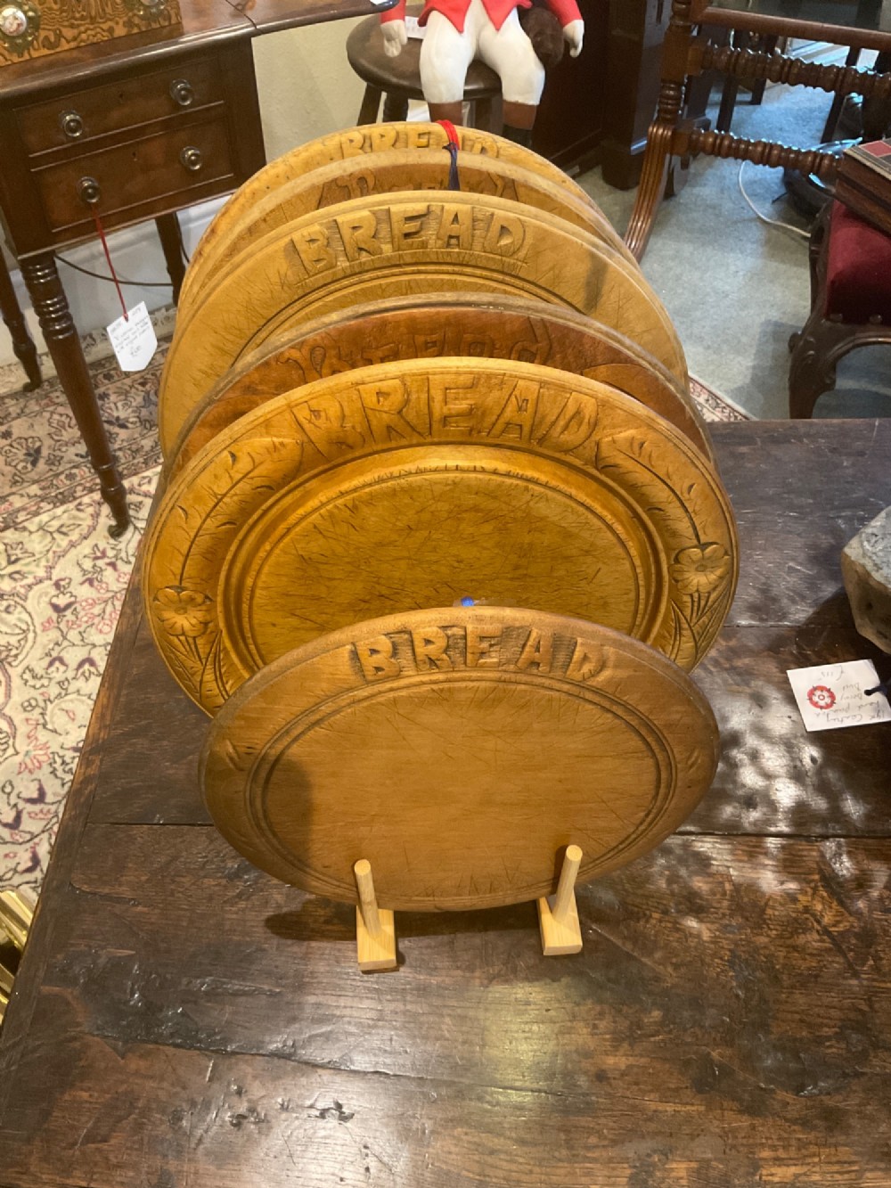 A circular antique wooden bread board featuring hand-carved lettering reading BREAD along the upper rim and knife marks across the centre. View V