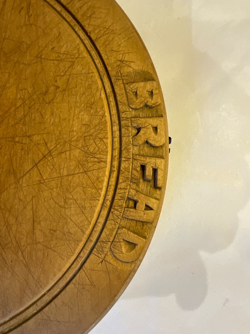 A circular carved wooden bread board with carved lettering spelling BREAD along the outer rim and visible knife marks. View II
